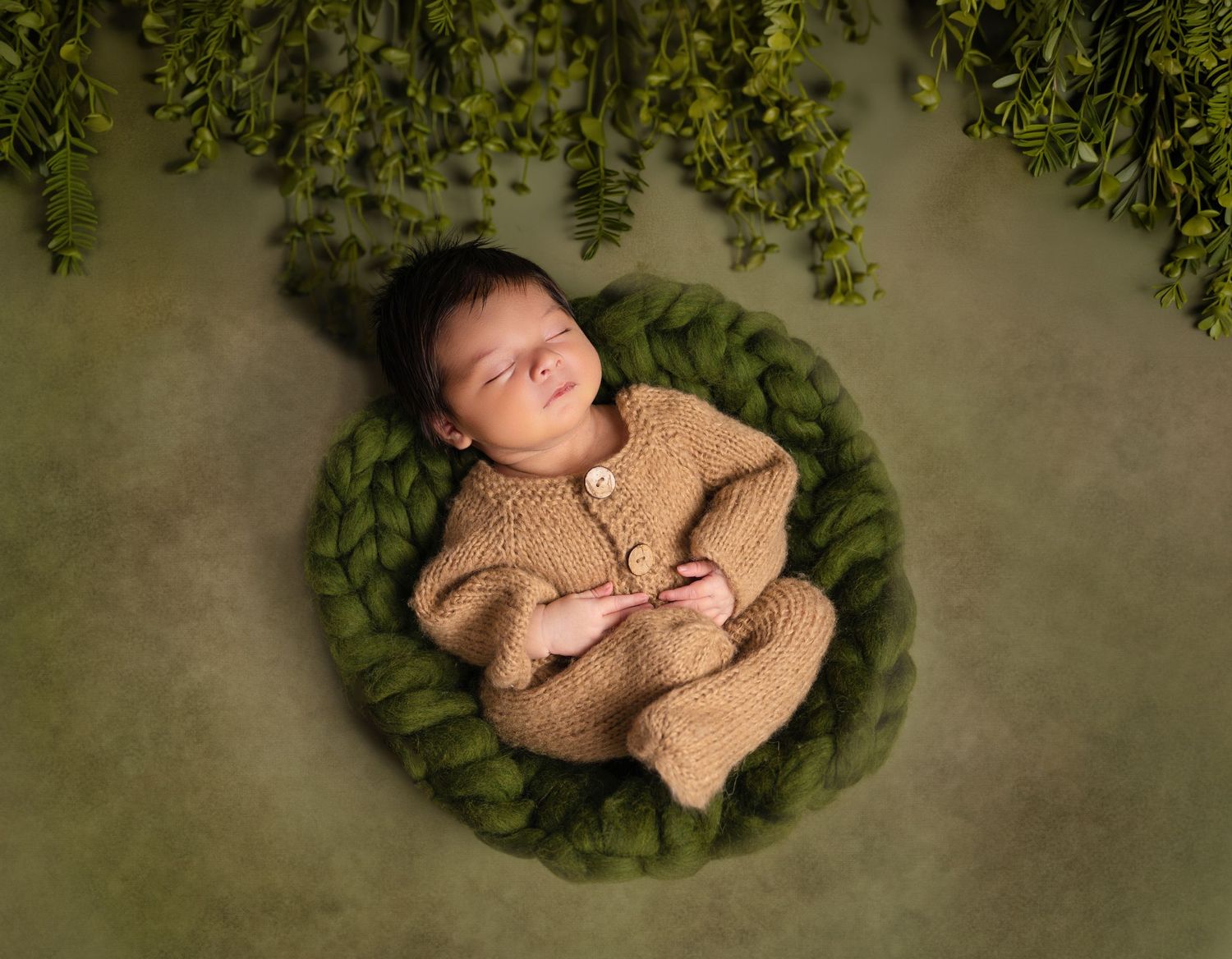 Sleeping newborn baby in tan knit romper nestled in green chunky yarn bowl surrounded by lush greenery.