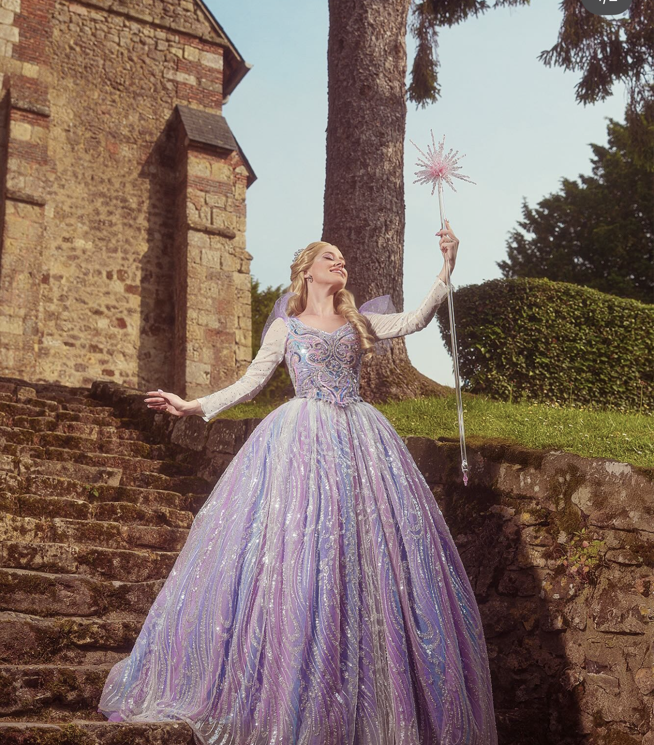 Woman in iridescent purple ballgown holding star wand on stone steps, fairytale princess costume photography.