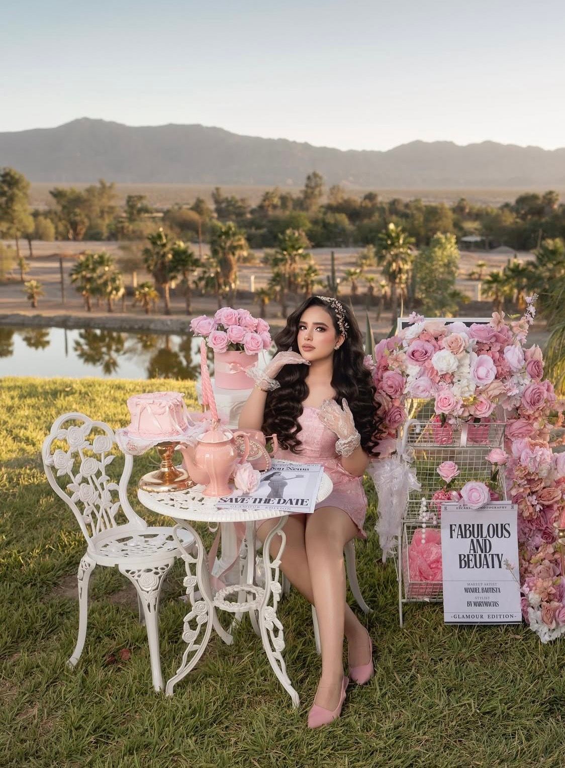 Elegant woman in pink dress at white garden table with floral decor and mountain backdrop at golden hour.