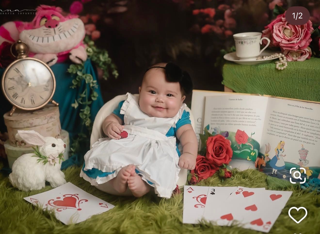 Smiling baby in an Alice in Wonderland costume sits on grass beside playing cards, a white rabbit plush, a pocket watch, and a storybook.