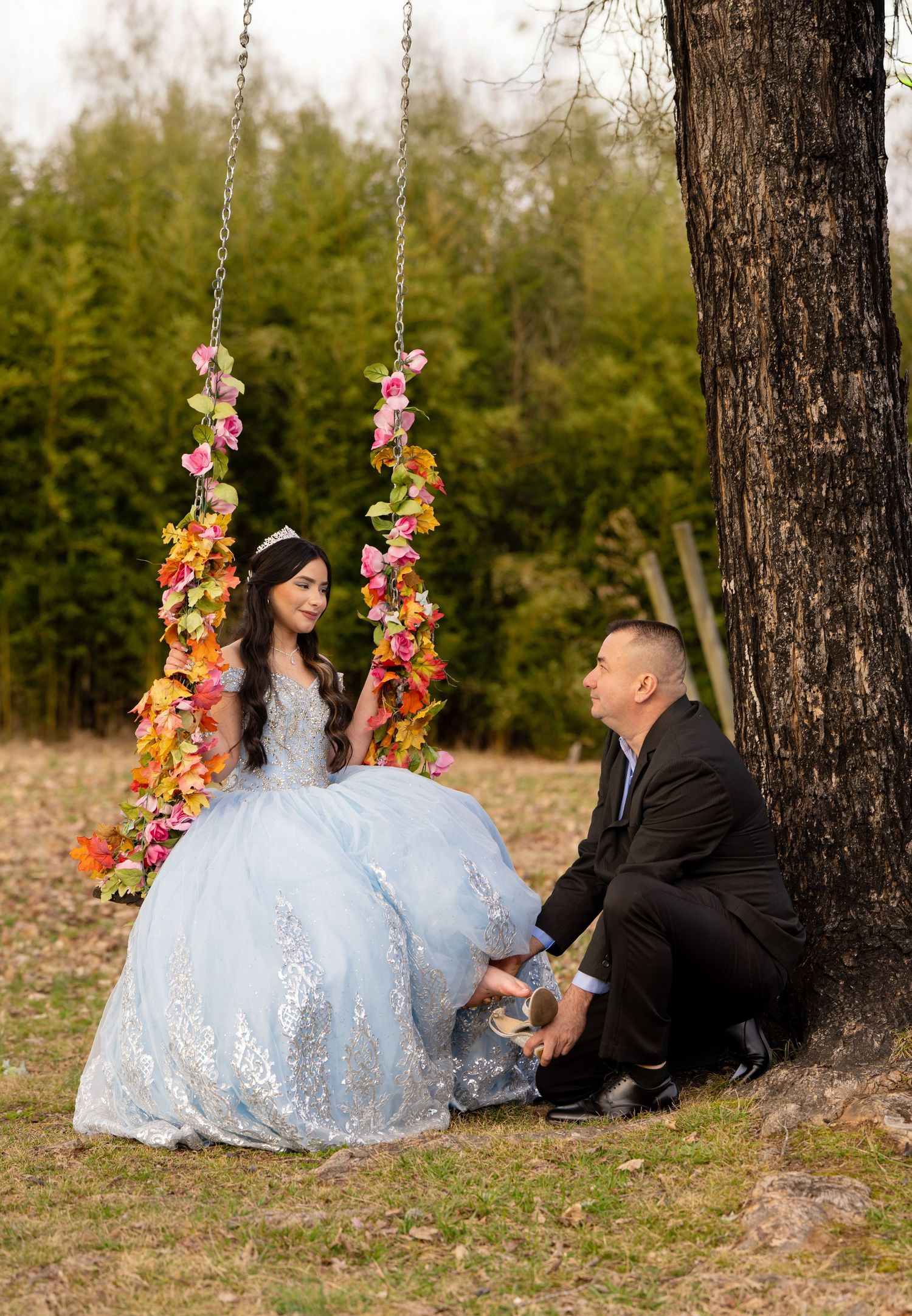 A girl in a blue ball gown sits on a floral swing while a man in a suit kneels beside her outdoors.