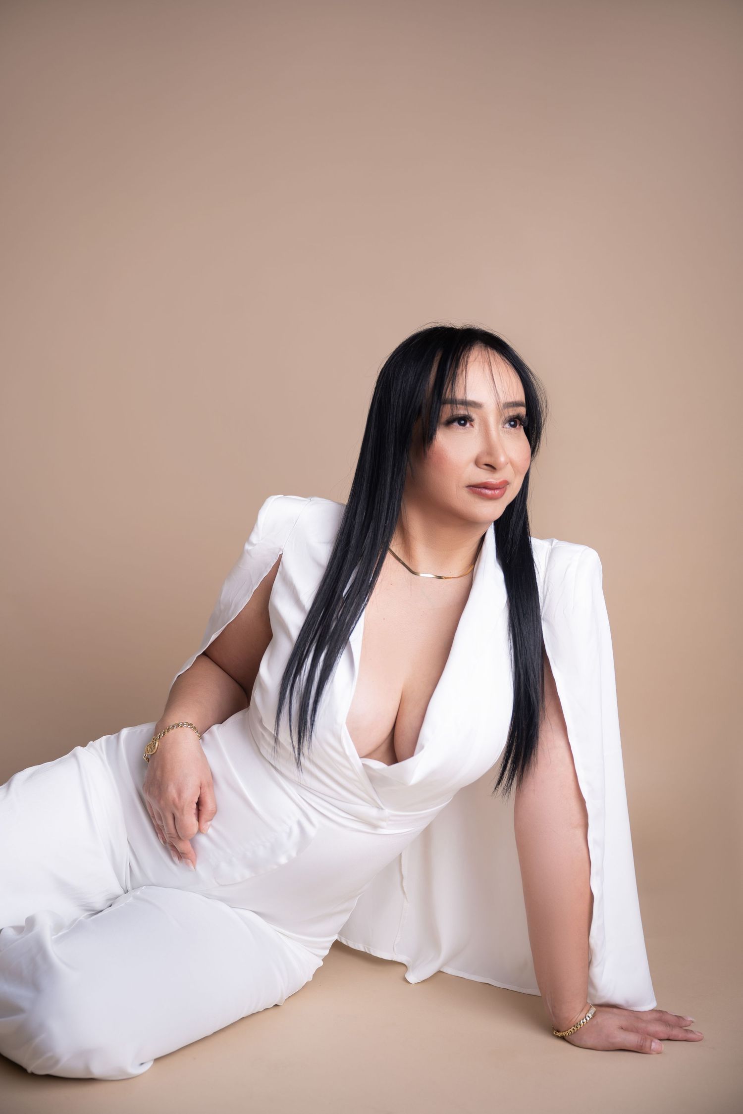 Woman with long black hair reclining on the floor in an elegant white caped jumpsuit against a warm beige studio backdrop.