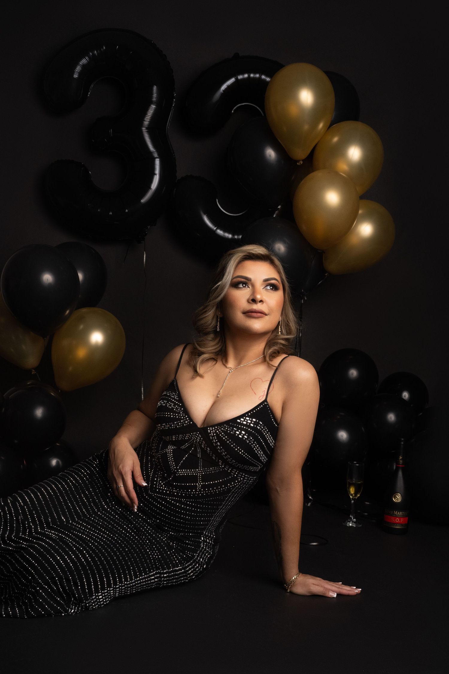 Woman in a sparkly black dress poses on the floor beside black and gold balloons spelling 33 for a glamorous birthday photoshoot.