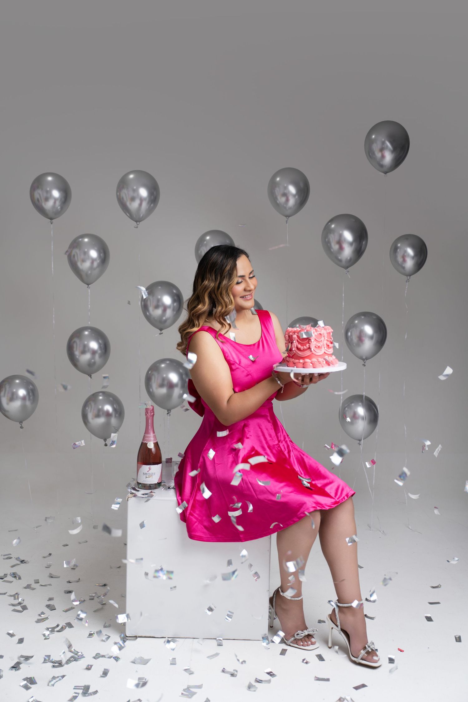 Woman in a hot pink dress sits on a white cube holding a pink cake, surrounded by silver balloons, confetti and a champagne bottle.