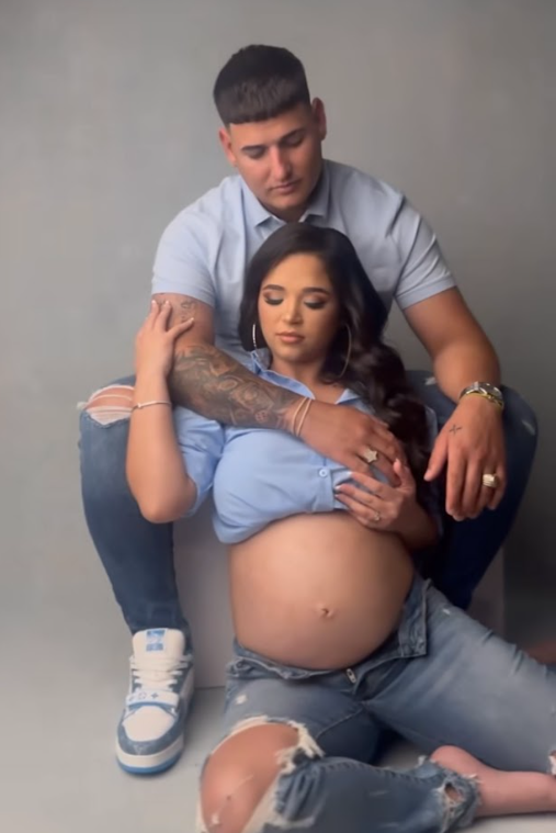 A tender maternity photo shows an expectant couple embracing in casual attire against a gray studio backdrop.