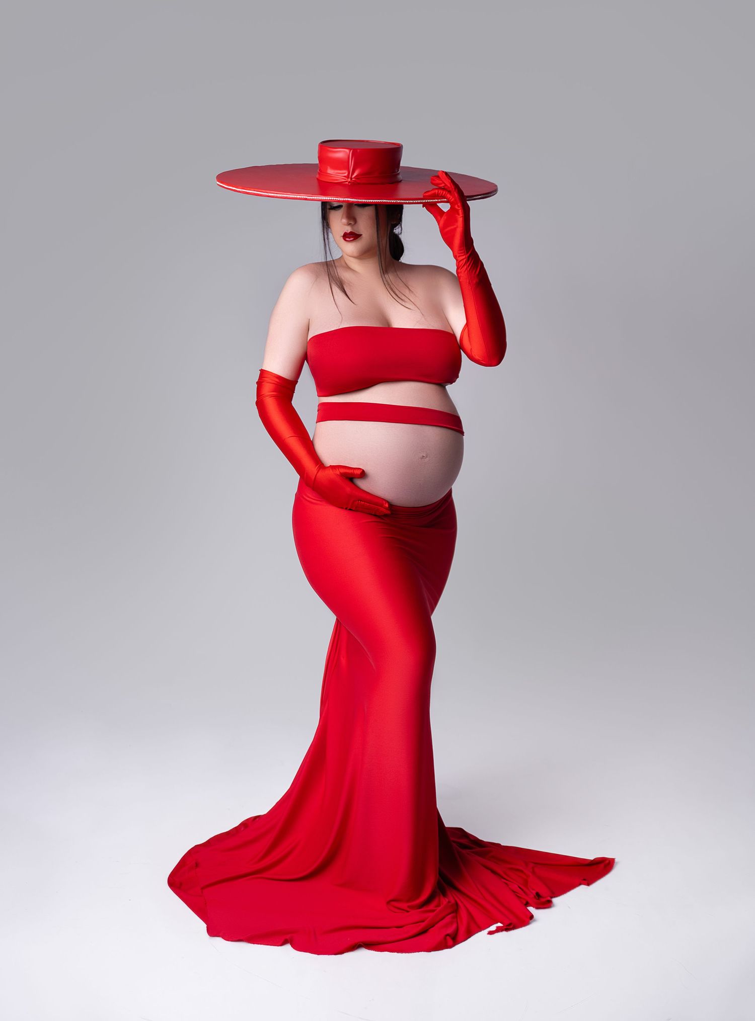 A figure in a flowing red gown and matching hat poses gracefully in a series of artistic maternity photos against a gray backdrop.
