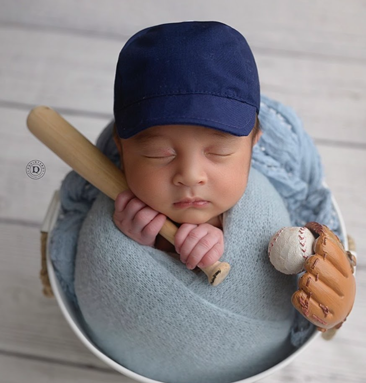 Sleeping baby in blue outfit holds a baseball bat and glove while resting on light blue cushion.