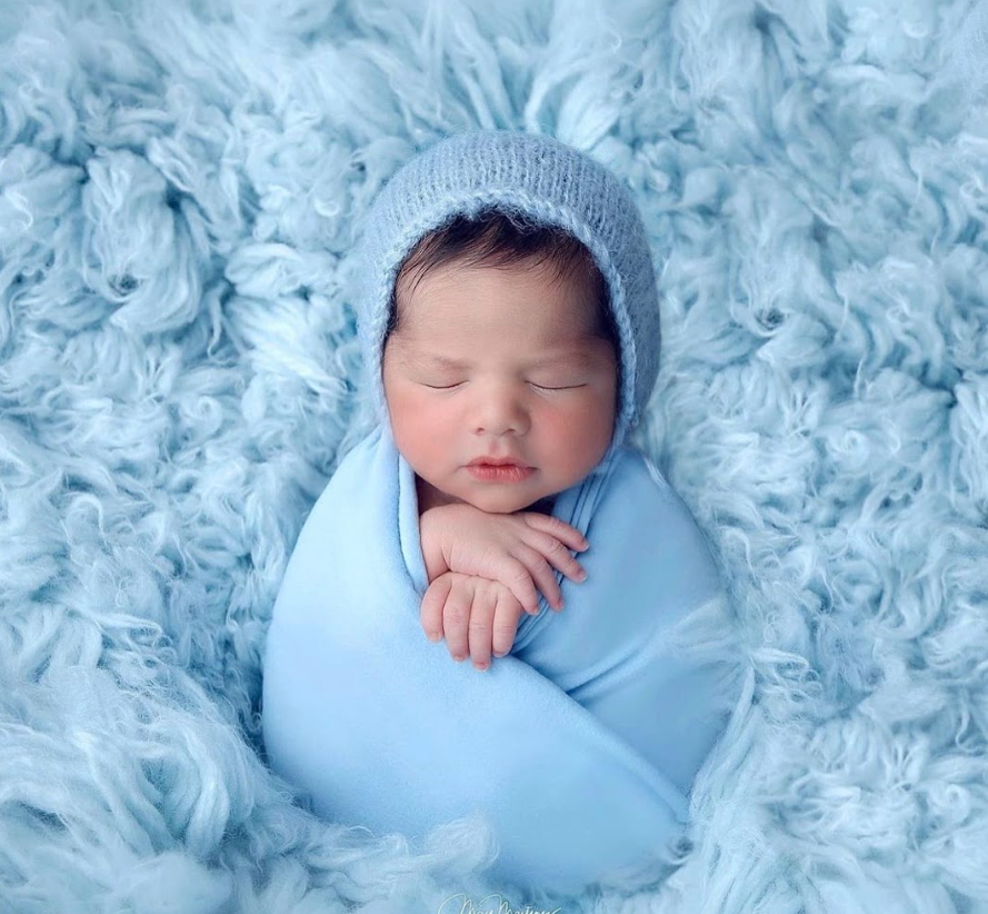 Sleeping newborn baby wrapped in soft blue blanket and knit hat on fluffy blue background.