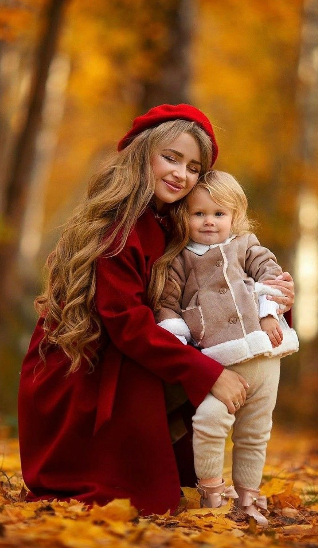 Loving parent in red cloak embraces small child among golden autumn leaves on ground during fall photoshoot.