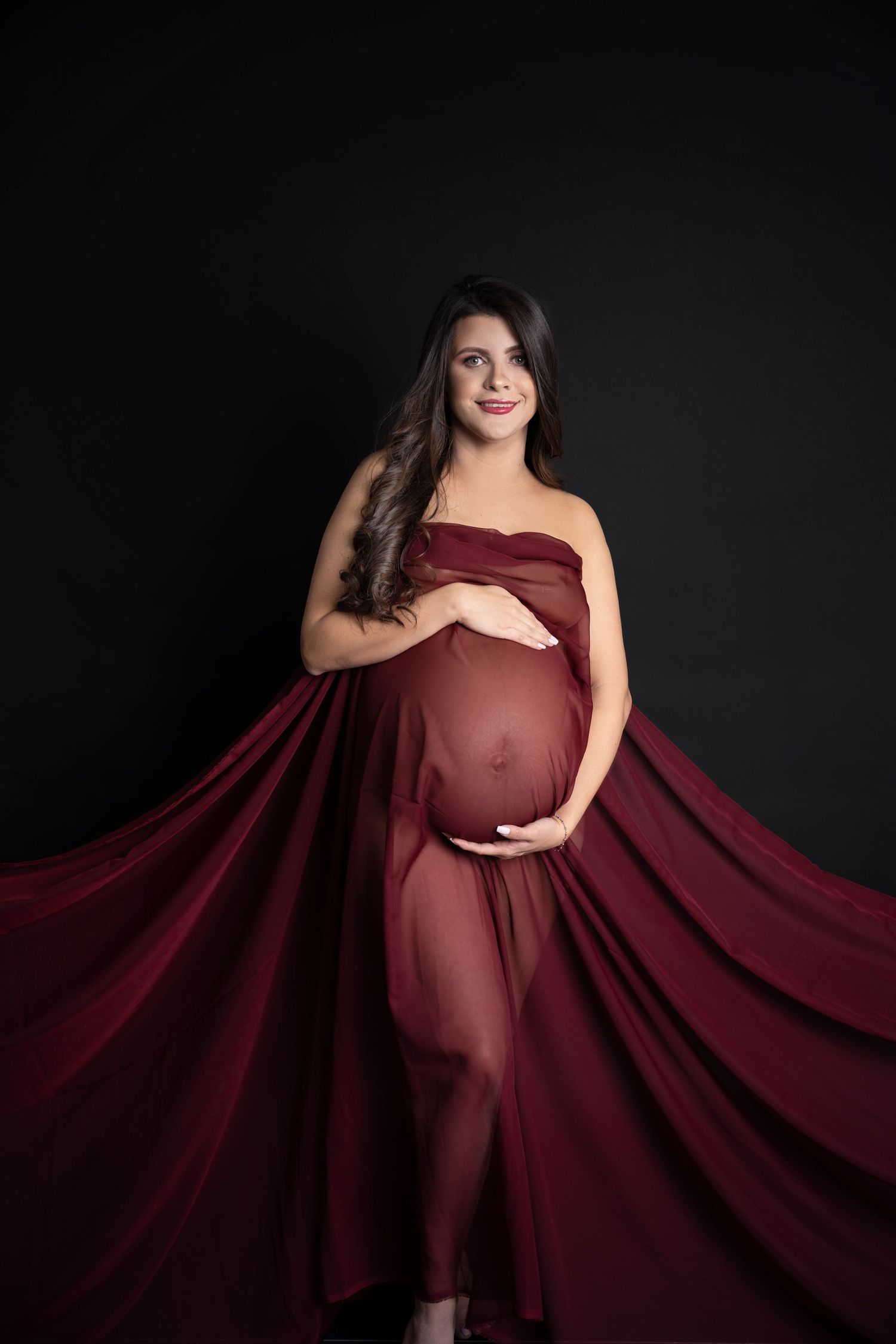 Pregnant woman in flowing red maternity gown cradling baby bump against dark studio background.