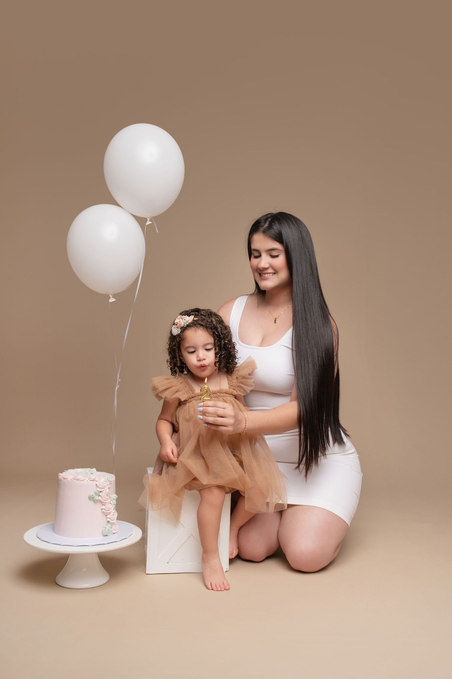 Mother and toddler daughter in matching white and tan outfits celebrate first birthday with white cake and balloons.