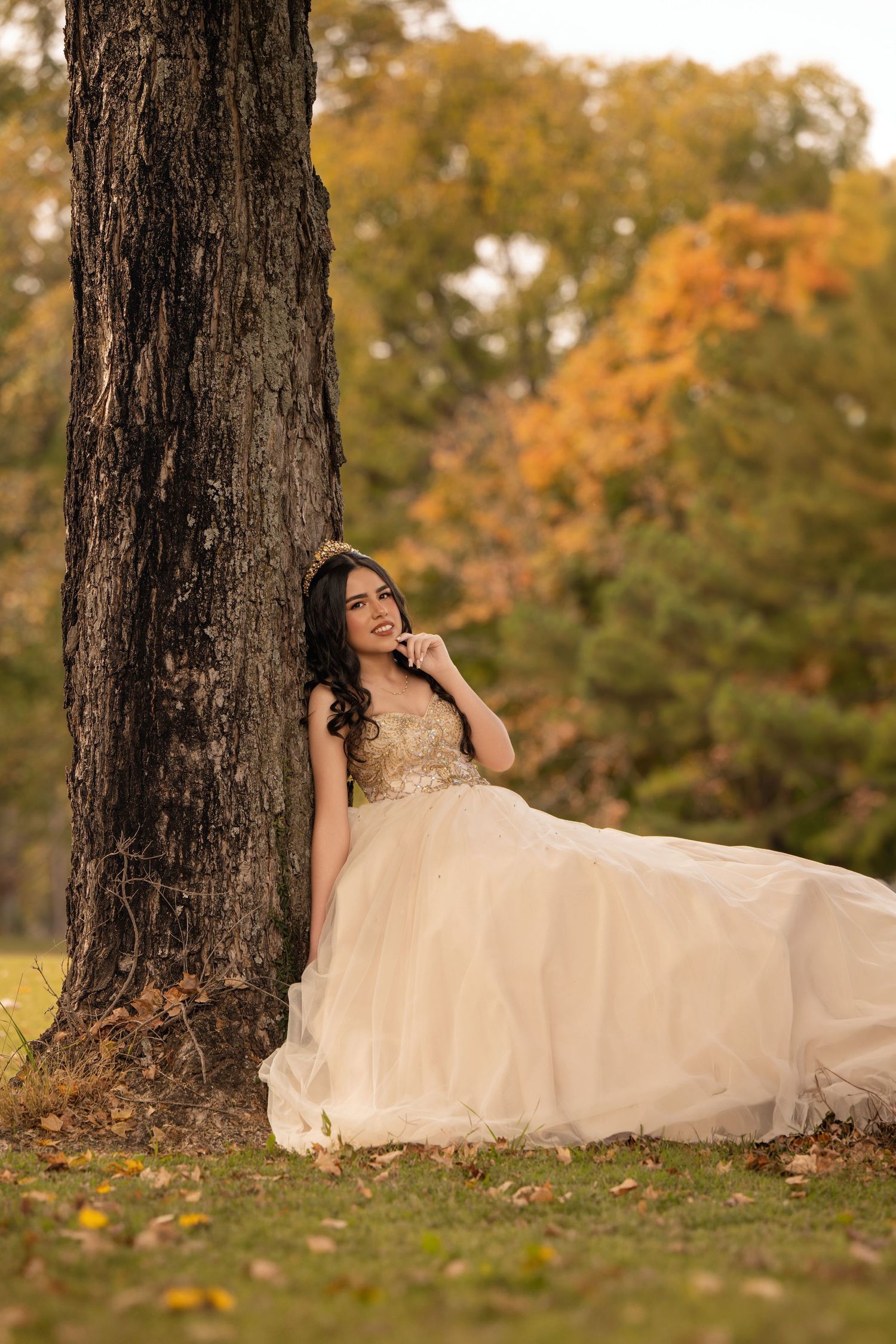 Young woman in elegant champagne ball gown leaning against tree in autumn park, wearing gold crown, smiling dreamily.