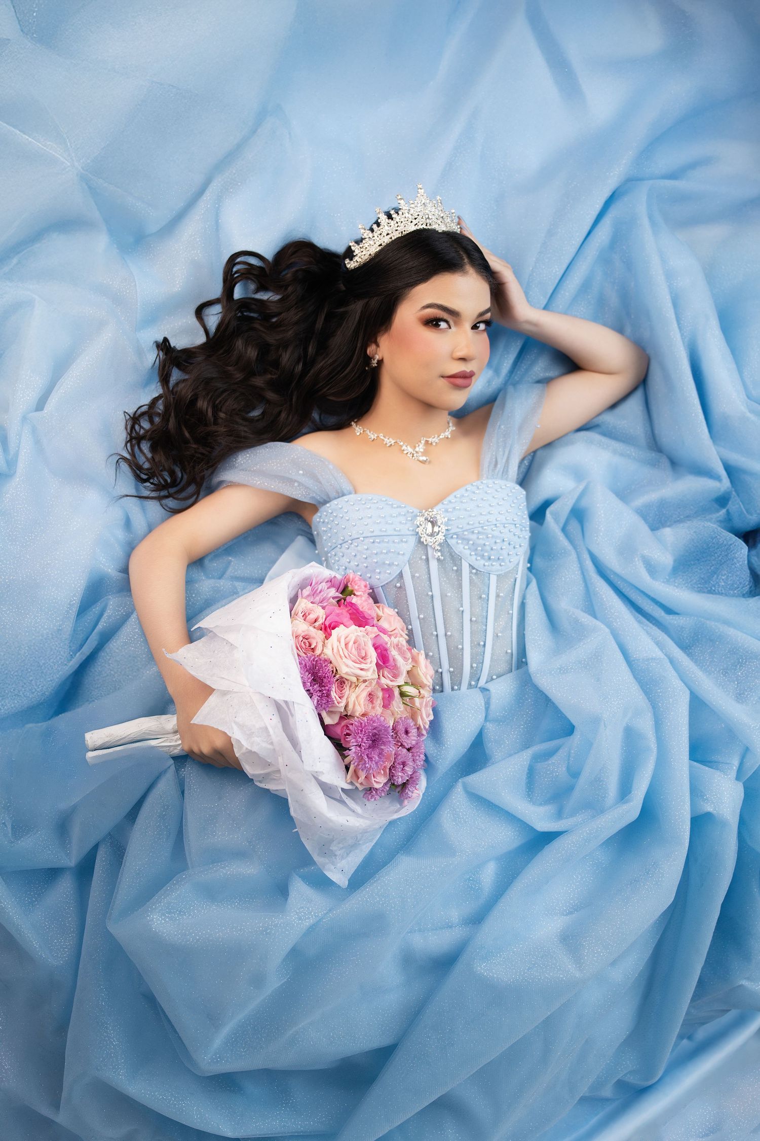 Young woman in blue ball gown and tiara holds pink bouquet, lying on flowing blue fabric.