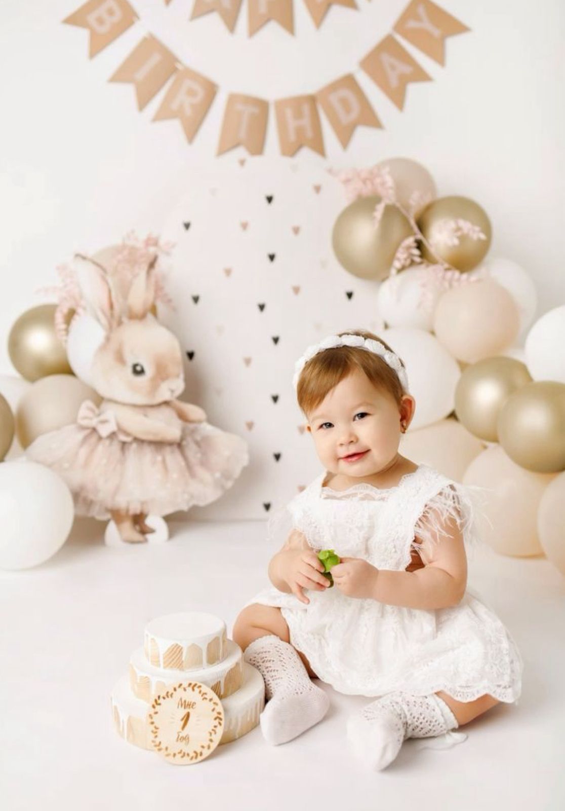 Soft pink and gold first birthday party setup with balloons, banners, and a small child in a white lace dress.