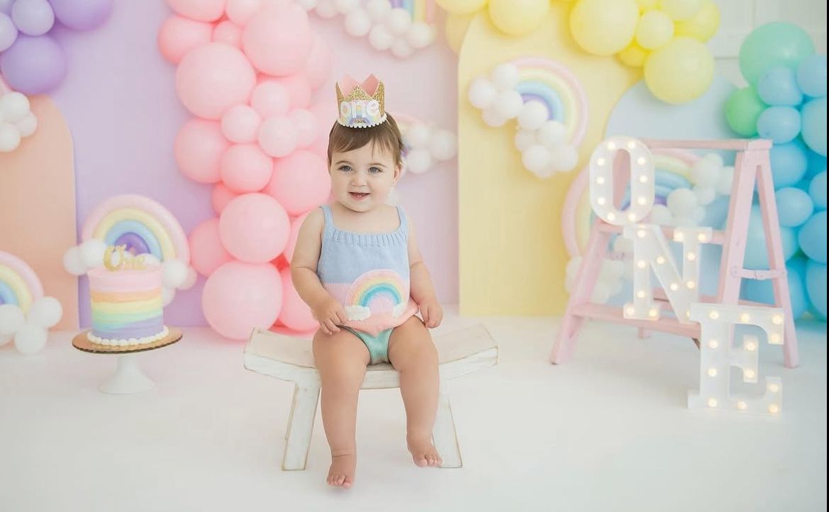 A cake smash photo session featuring a pastel balloon arch and rainbow-themed decorations for a first birthday celebration.
