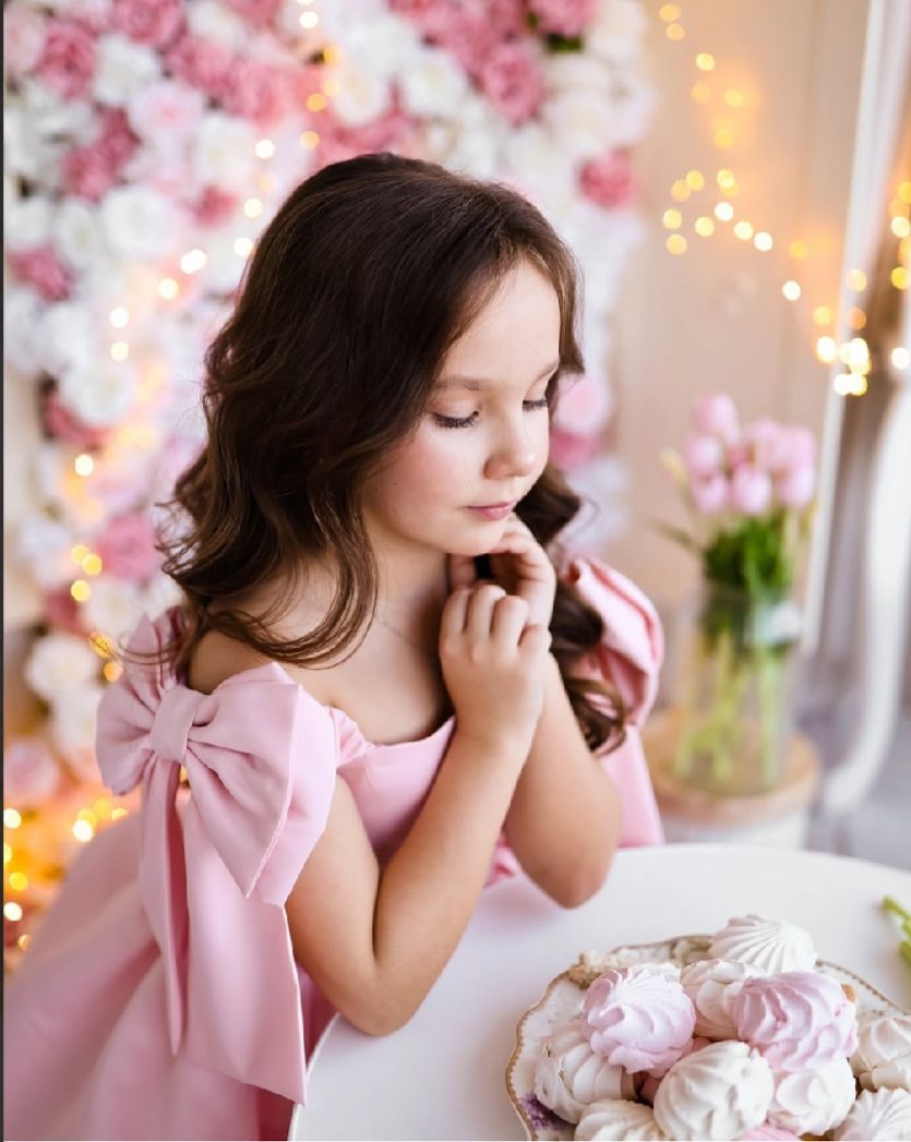 Dreamy portrait in pink dress against floral backdrop with fairy lights and pastel decorations.