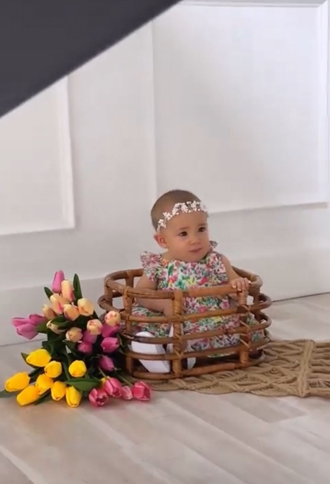 Baby in floral dress sits in decorative wooden basket next to colorful tulip bouquet during spring photo session.