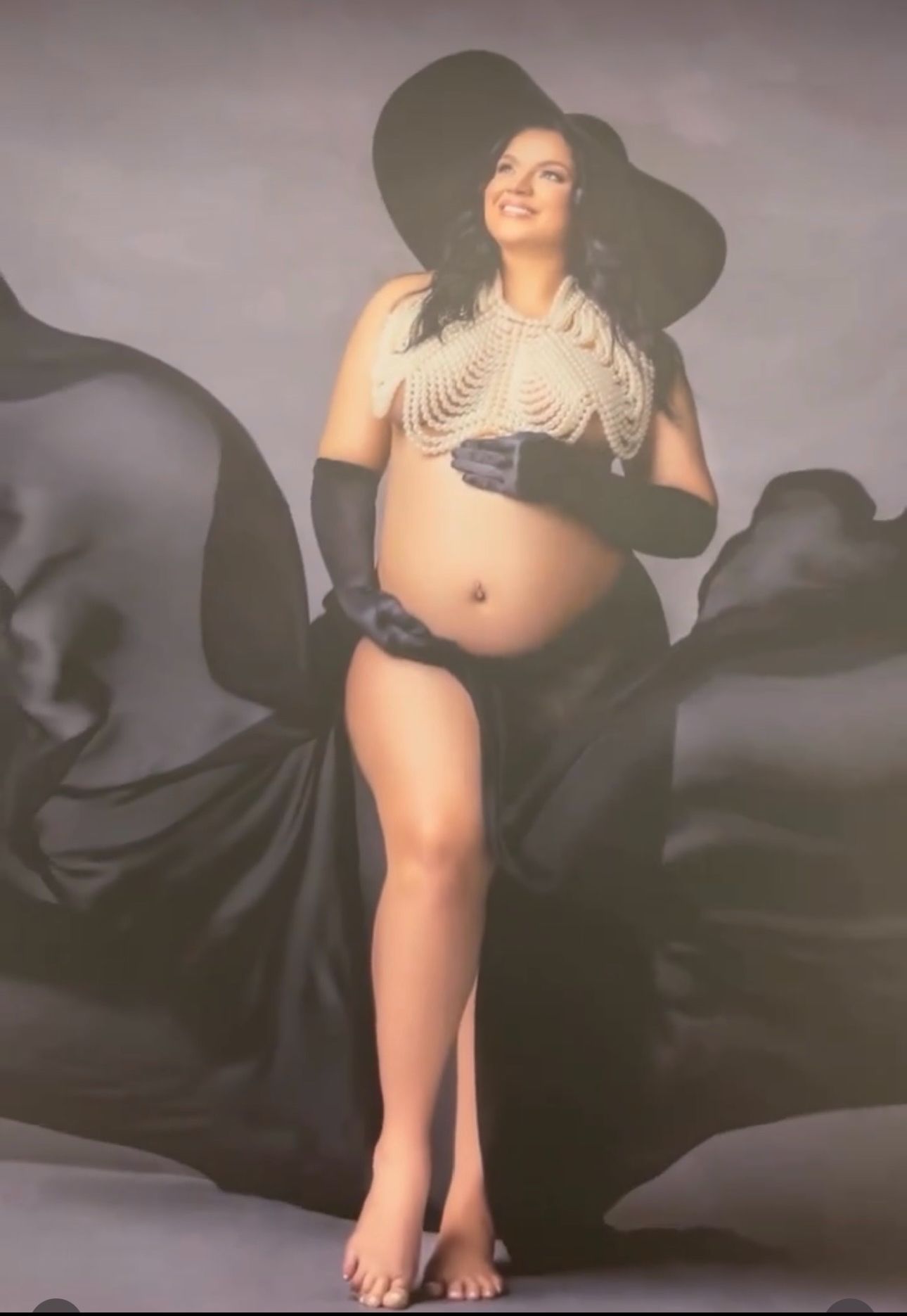 Pregnant woman in elegant black hat and gloves with pearl body jewelry, posing with flowing black fabric backdrop.