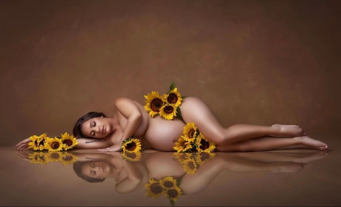 Pregnant woman lying gracefully on floor surrounded by sunflowers in artistic maternity photography session.
