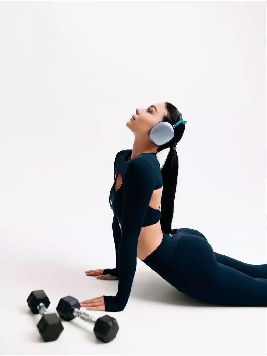 Woman in dark athletic wear and headphones poses on floor beside dumbbells against white background.