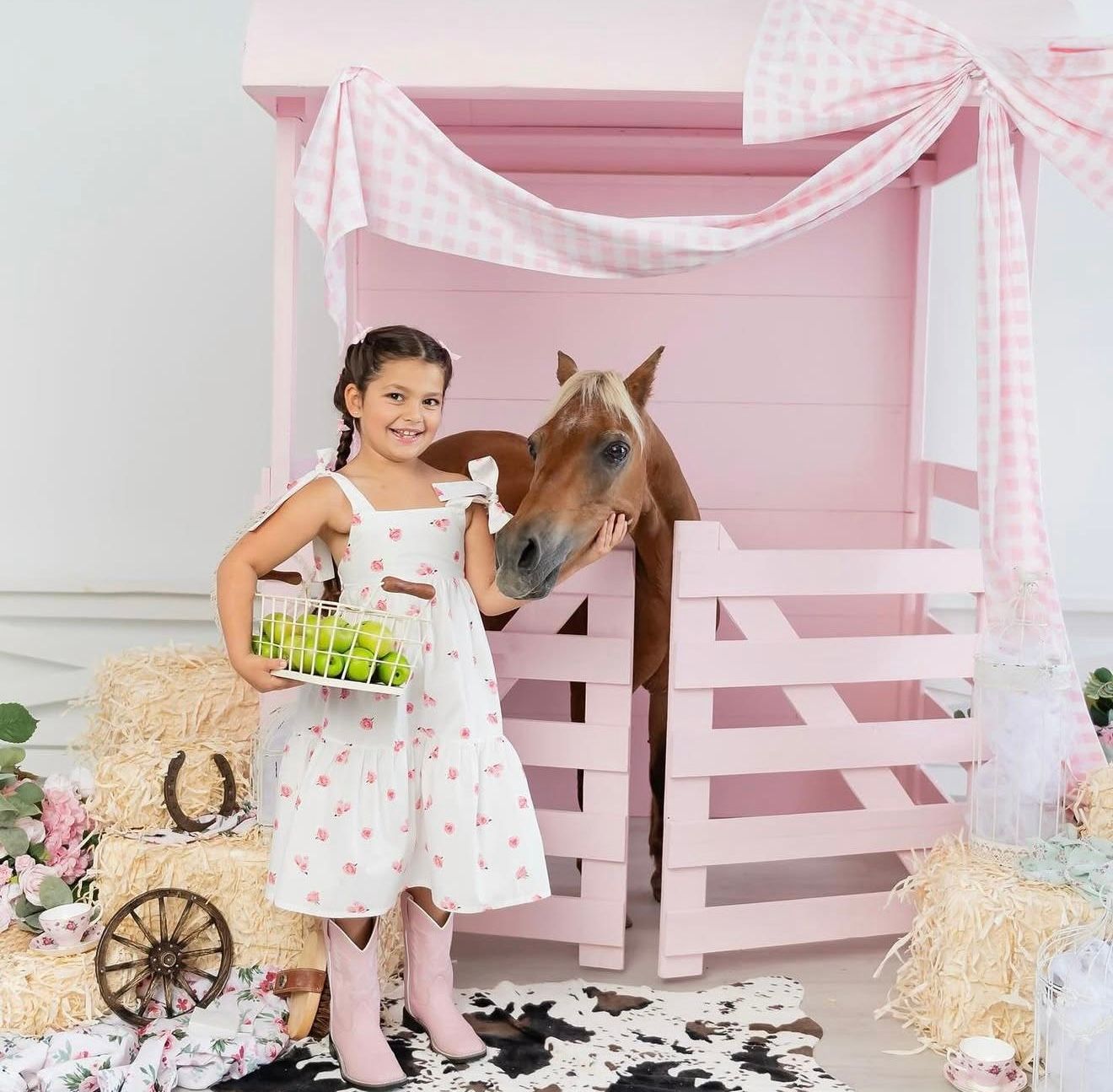 Young girl in white floral dress and pink boots holds basket near a brown horse in a pink barn-themed photo setting.