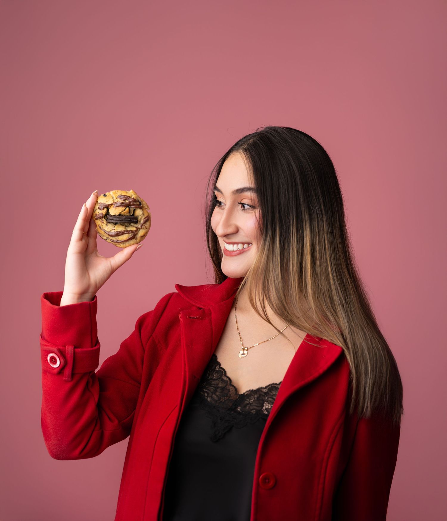 A person in a red coat holding up a hamburger while smiling against a pink background.