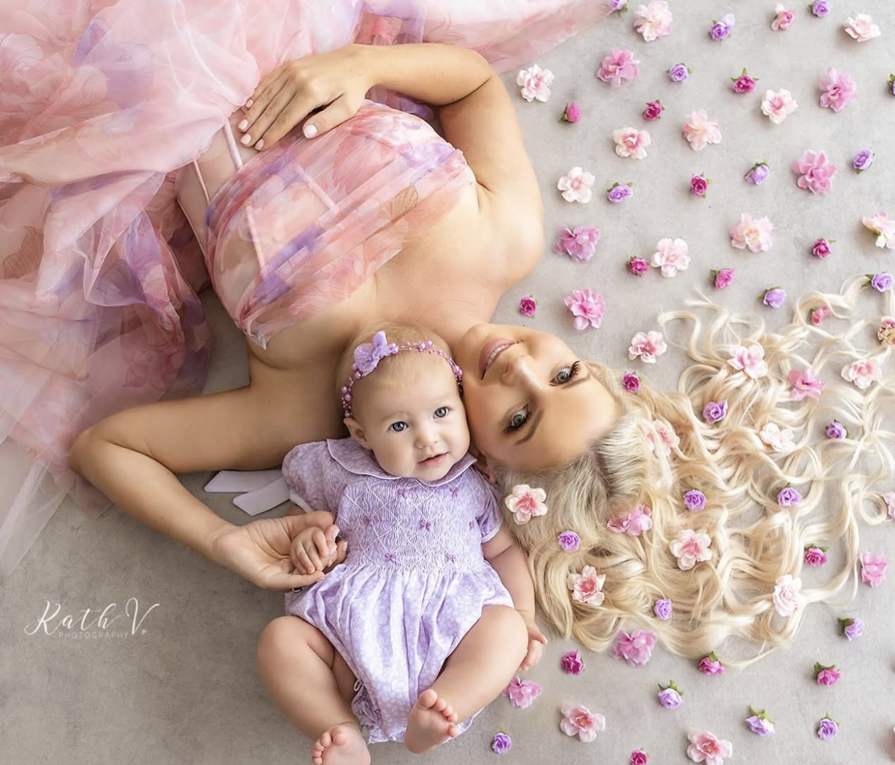 Mother and baby girl lying on floor surrounded by pink flowers, wearing matching lavender and pink outfits in studio photo.