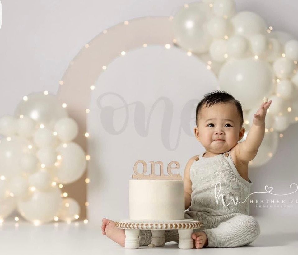 Baby's first birthday cake smash photo with white balloons, fairy lights, and a white cake with “one” topper.