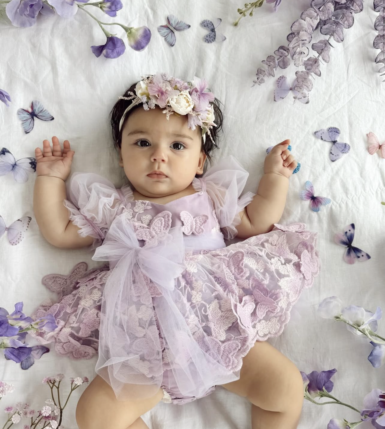 Baby girl in purple lace dress and floral headband lying on butterfly-printed white blanket.