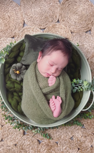 A newborn baby swaddled in olive green fabric sleeps peacefully in a vintage enamel basin surrounded by greenery.