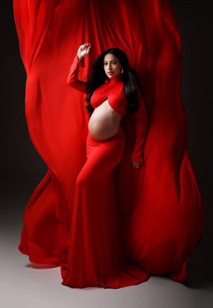 Dramatic maternity photo with flowing red fabric and dress against dark background creating ethereal movement.