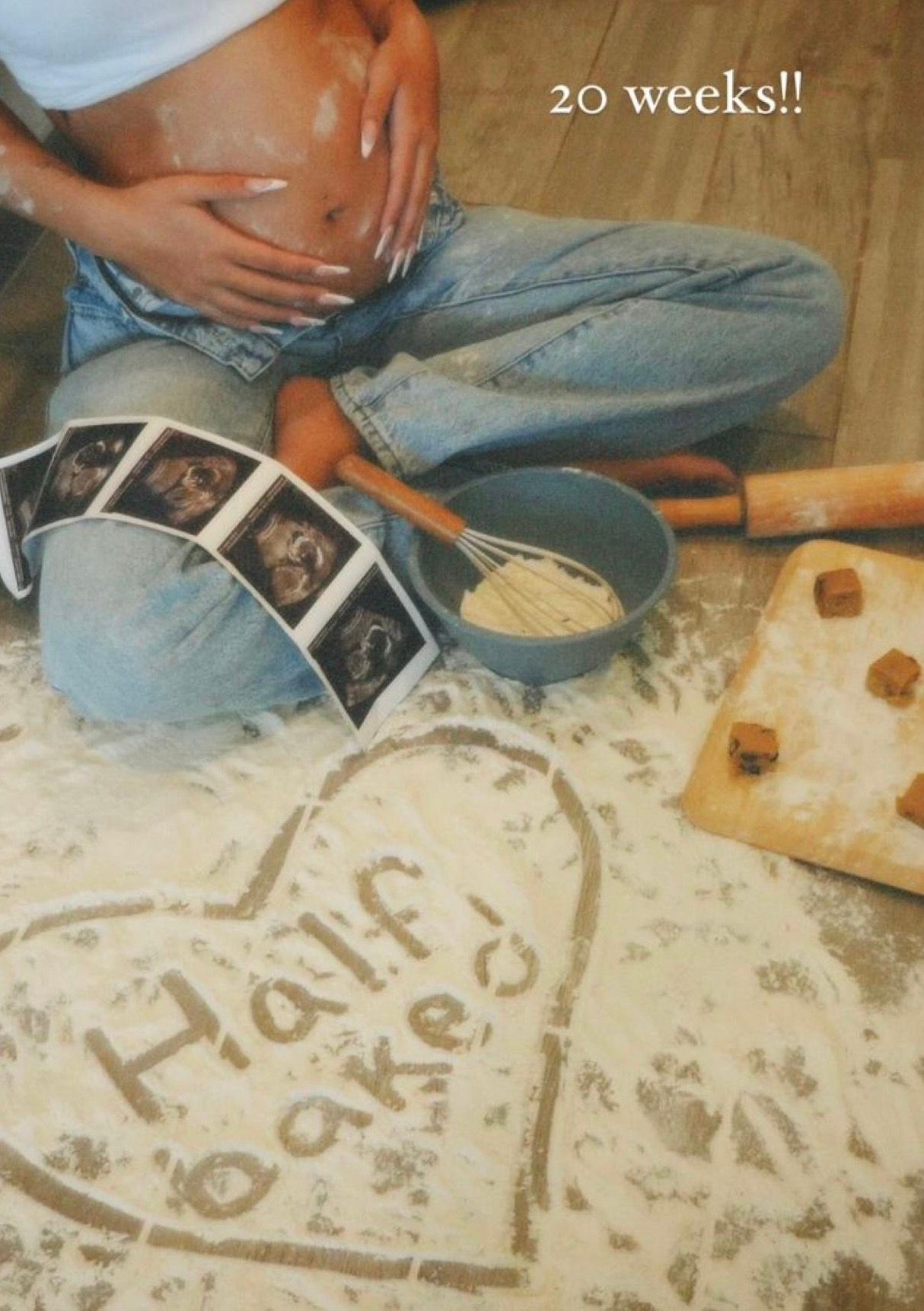 Pregnant woman sitting cross-legged with flour spelling Half Baked, ultrasound photos, and baking tools at 20 weeks.