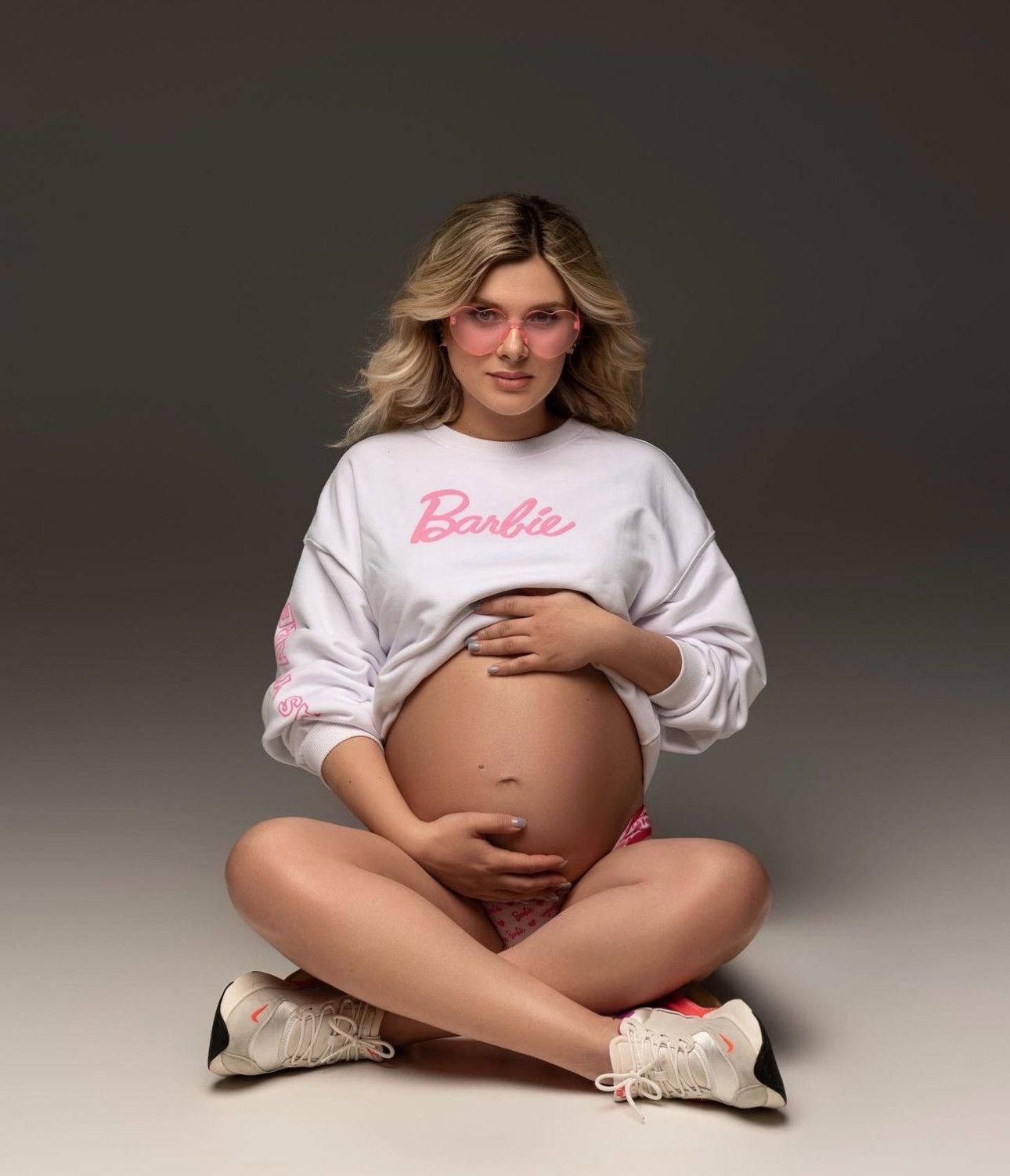 Pregnant woman sitting cross-legged wearing a white Barbie sweatshirt, cradling her baby bump, with pink sunglasses.