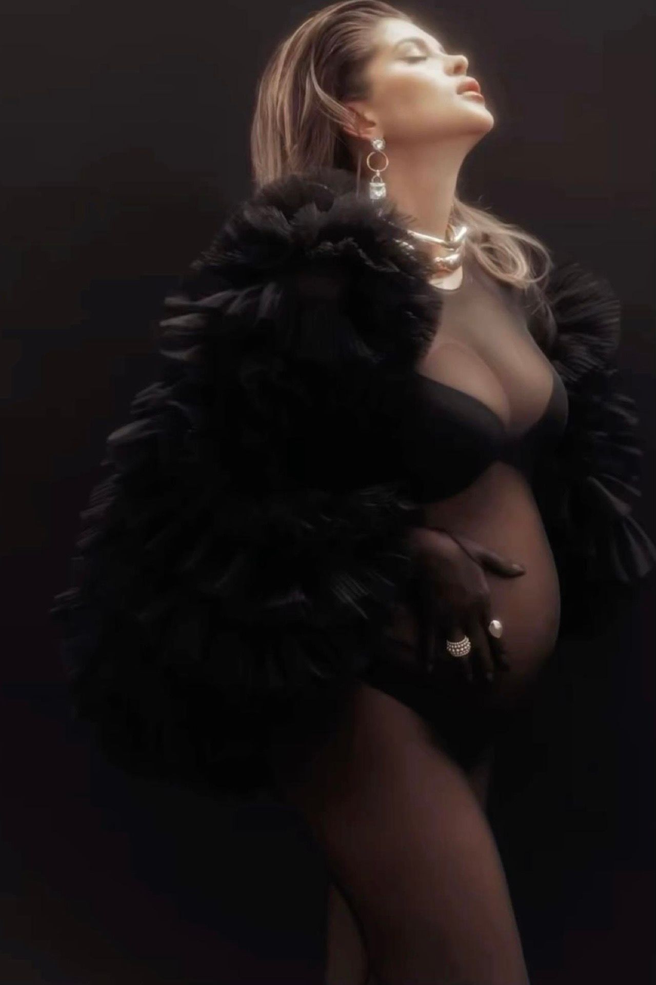 Pregnant woman in black feather coat and lingerie poses gracefully with pearl jewelry in a dramatic dark studio setting.
