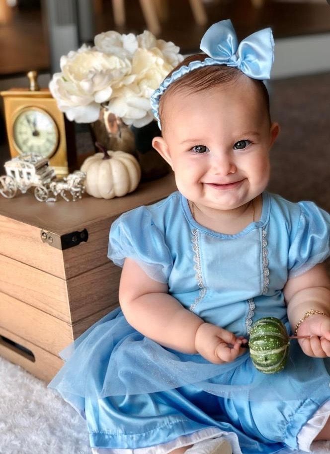 Smiling baby girl in blue Cinderella dress with bow headband sitting near white pumpkin and floral decor.