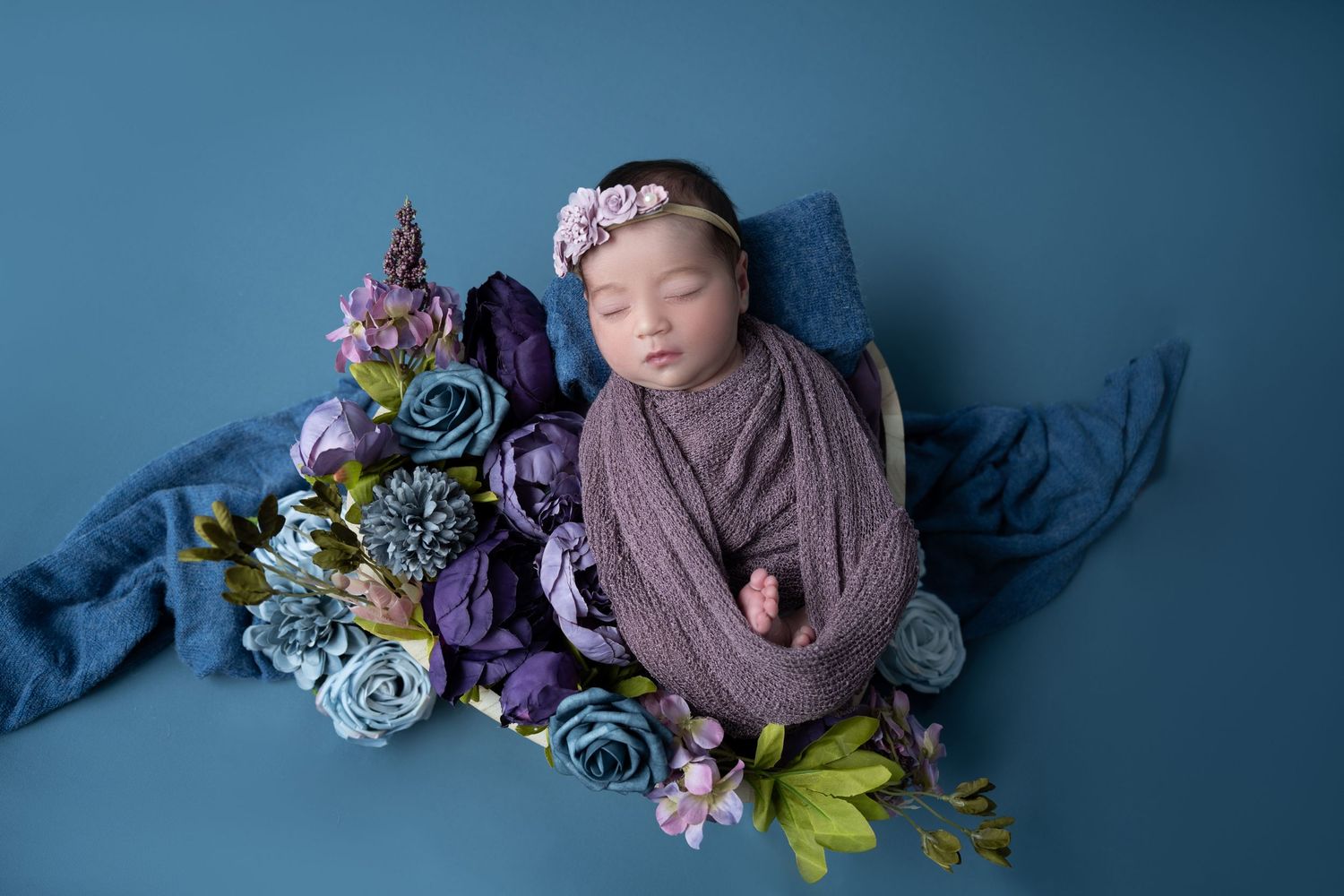 Sleeping newborn baby wrapped in purple swaddle with floral headband surrounded by blue and purple flowers on teal backdrop.