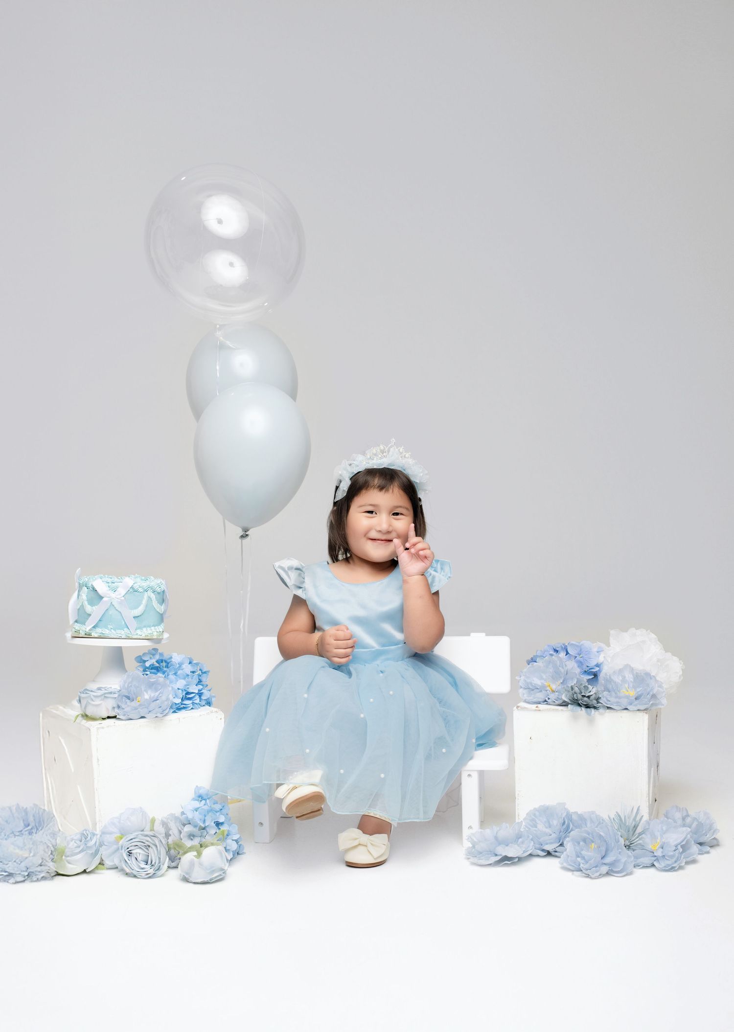 A smiling toddler girl in a blue tulle dress sits among balloons and floral decorations at a birthday celebration.