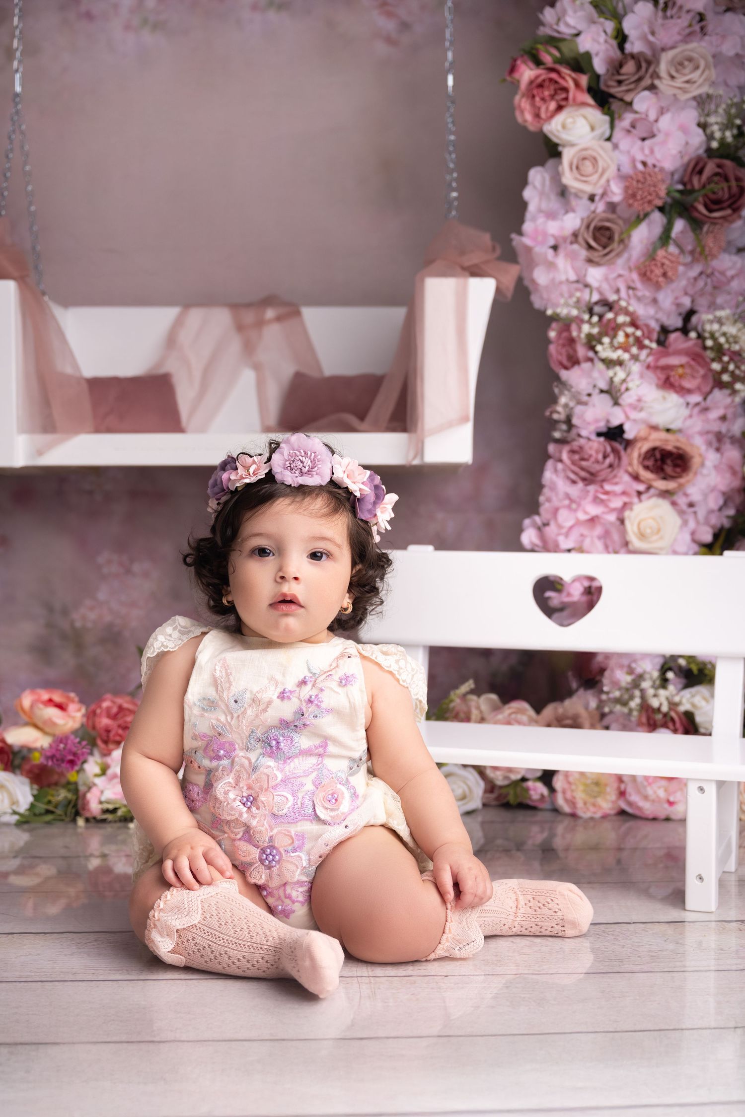 Adorable baby girl in floral romper and flower crown sits before a pink floral backdrop with a white bench.