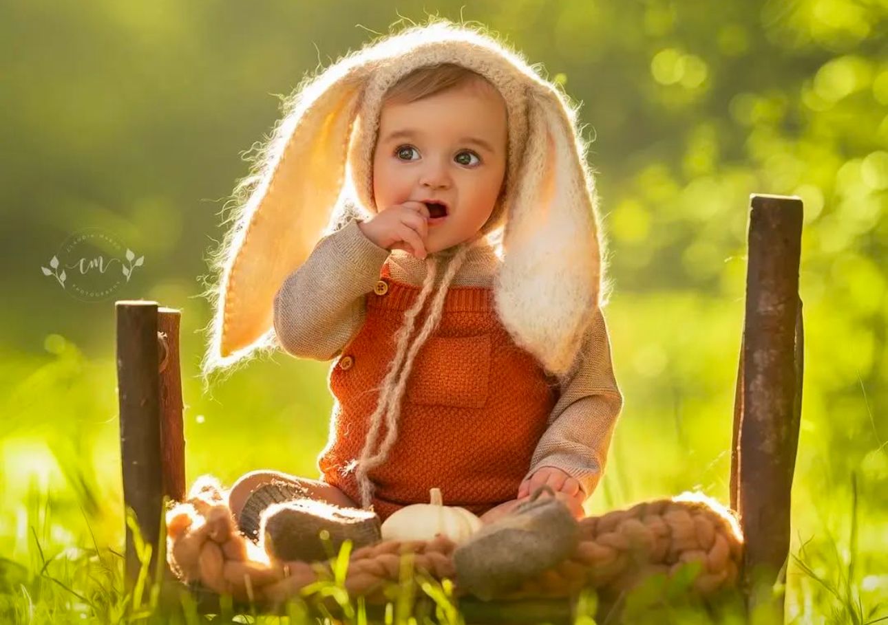 Adorable baby in orange bunny costume with floppy ears sitting in grass, chewing a carrot in sunny garden.