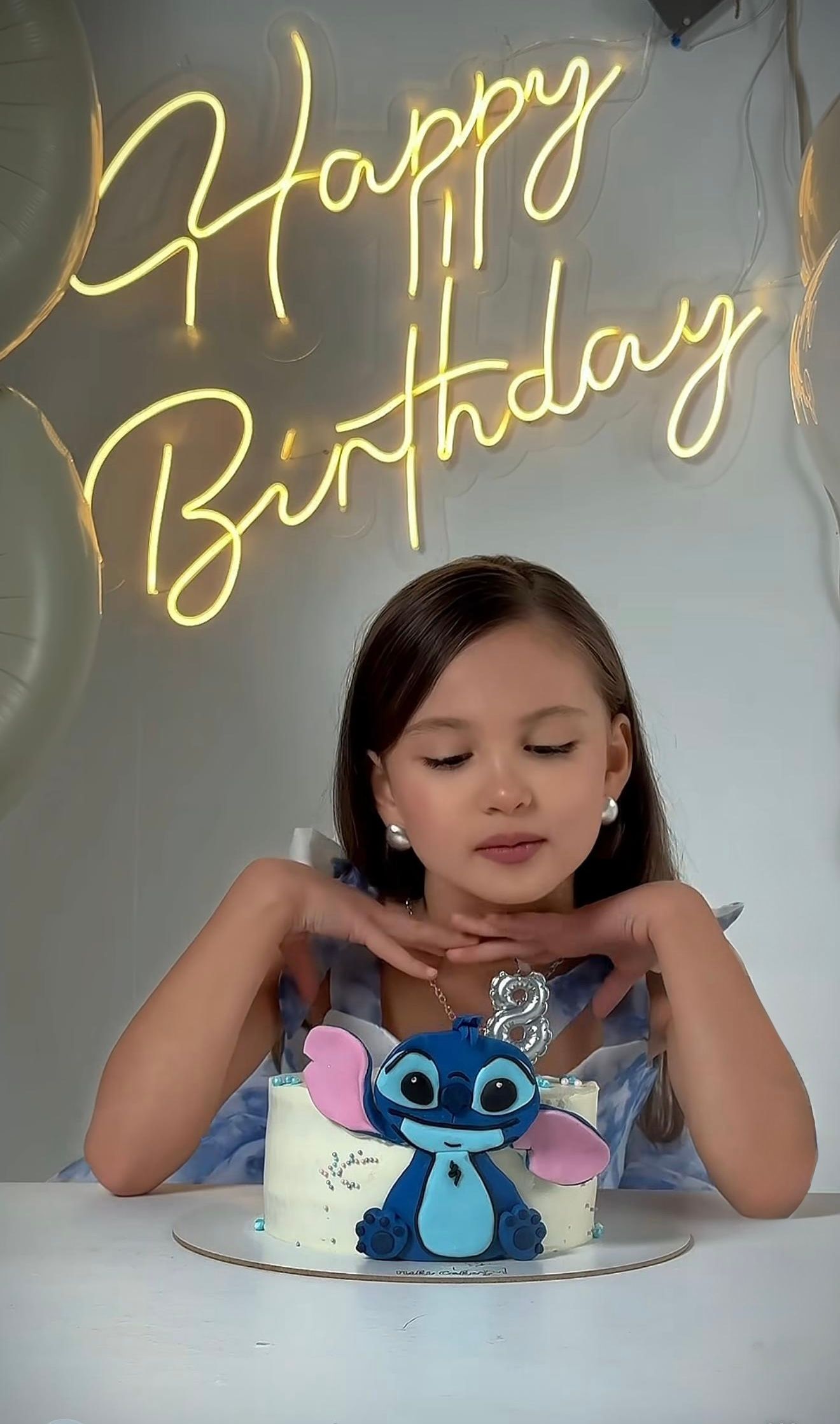 Young girl celebrating birthday with Stitch cake under Happy Birthday neon sign.