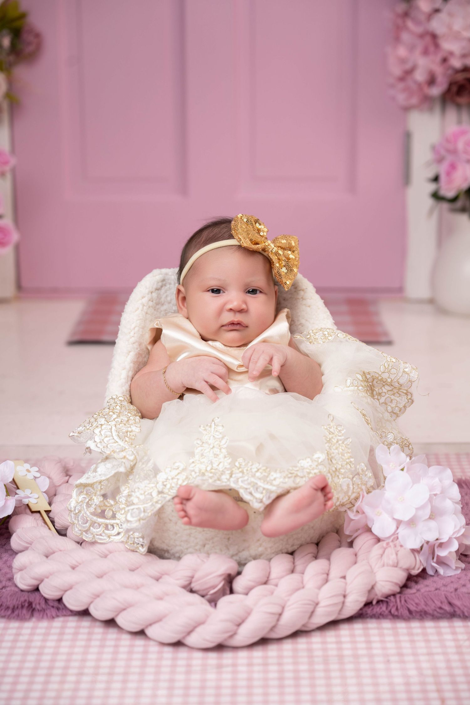 Newborn baby girl in white lace dress and gold bow headband sitting in fluffy basket with pink floral backdrop.