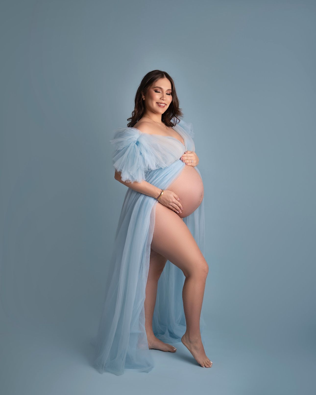Pregnant woman in sheer light blue tulle maternity gown posing gracefully against a soft blue studio background.