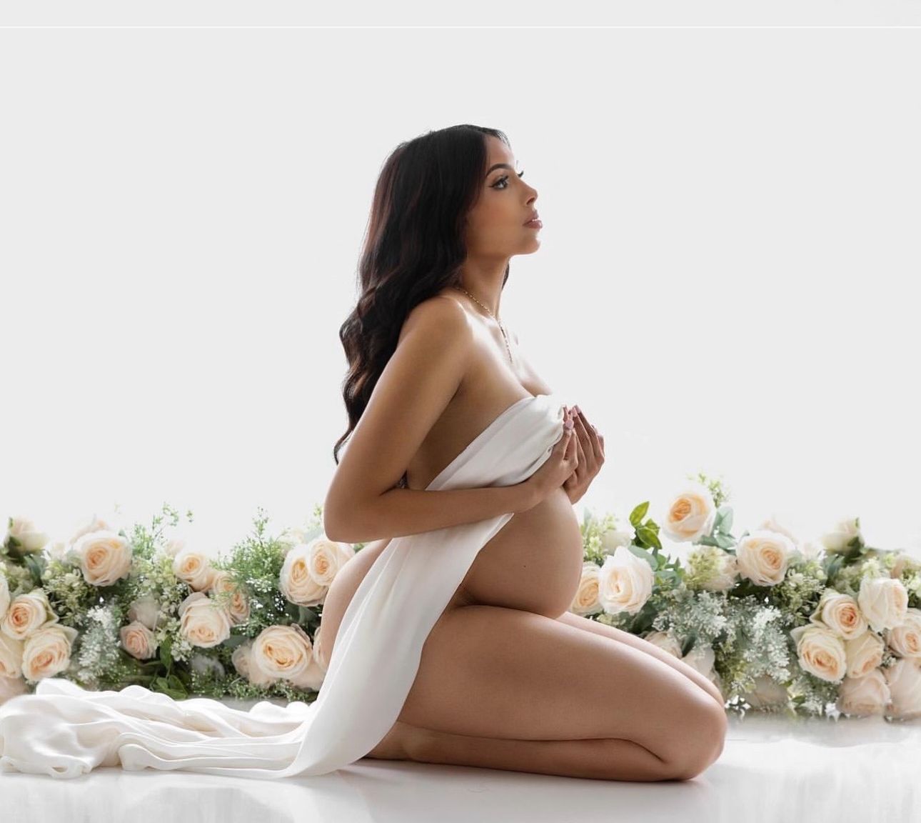 Pregnant woman kneeling gracefully draped in white fabric surrounded by soft peach roses in an elegant maternity portrait.