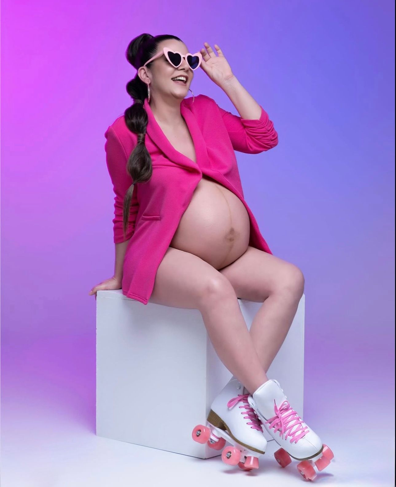 Pregnant woman in pink blazer and heart sunglasses sitting on white cube wearing roller skates on purple background.