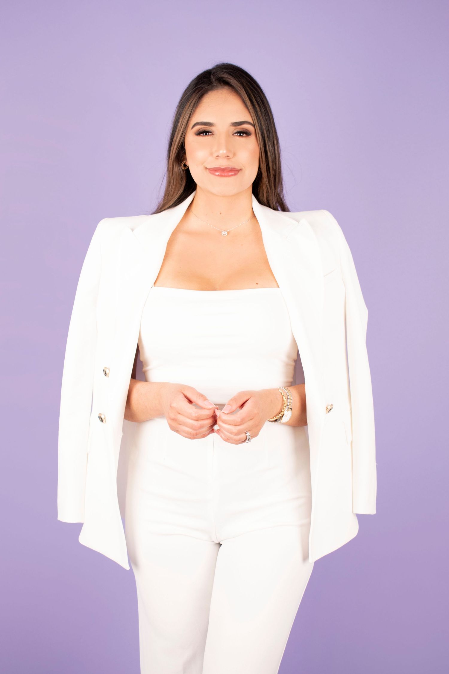 Woman in white blazer and pants poses confidently against purple background, wearing gold jewelry and watch.