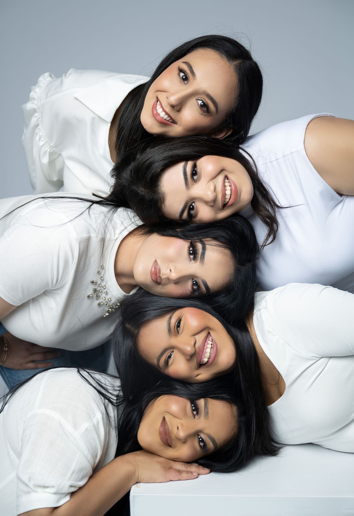Four young women with dark hair wearing white tops pose together stacked vertically, smiling at the camera.