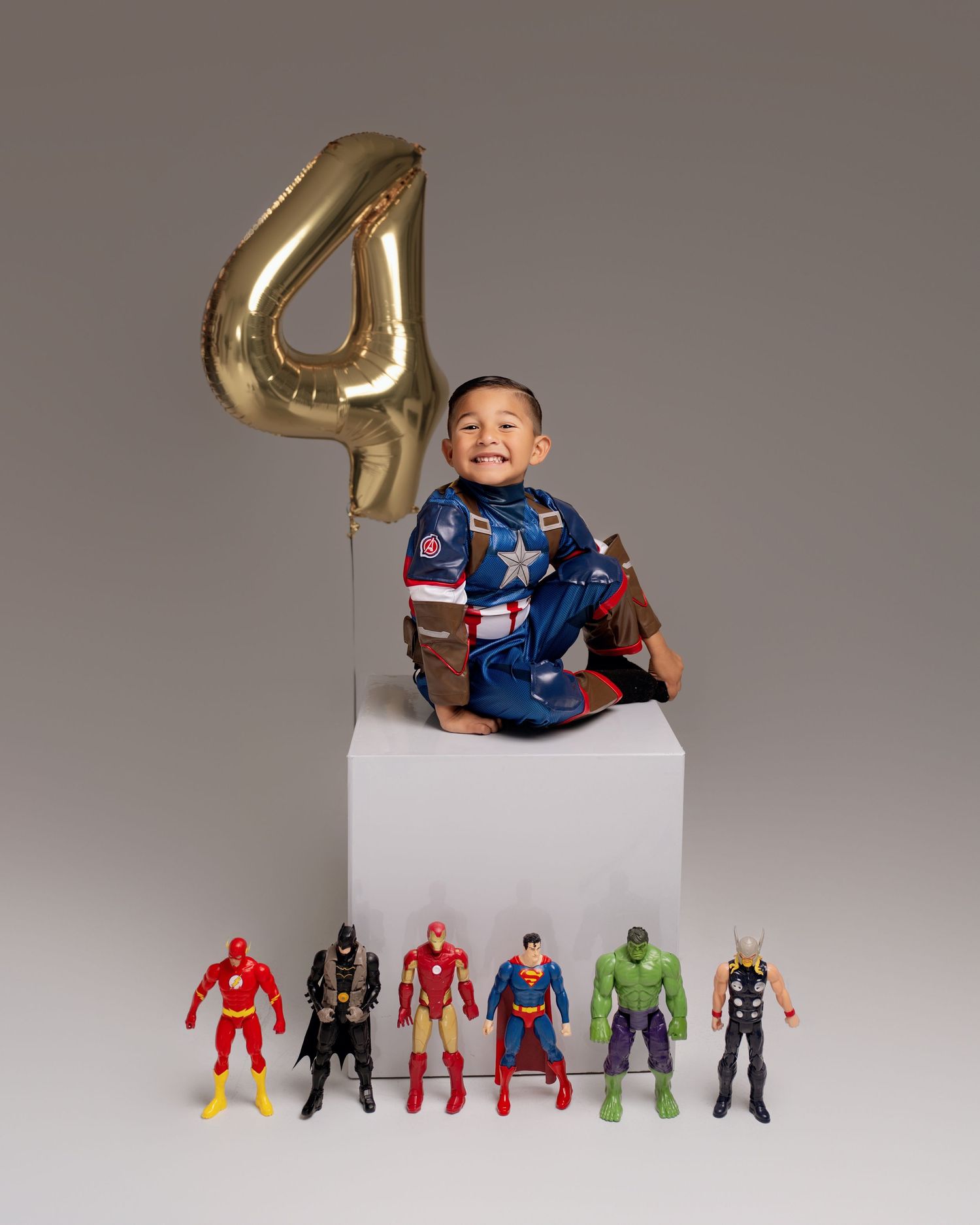 Boy in Captain America costume sits on white cube with superhero toys and gold number 4 balloon for 4th birthday.