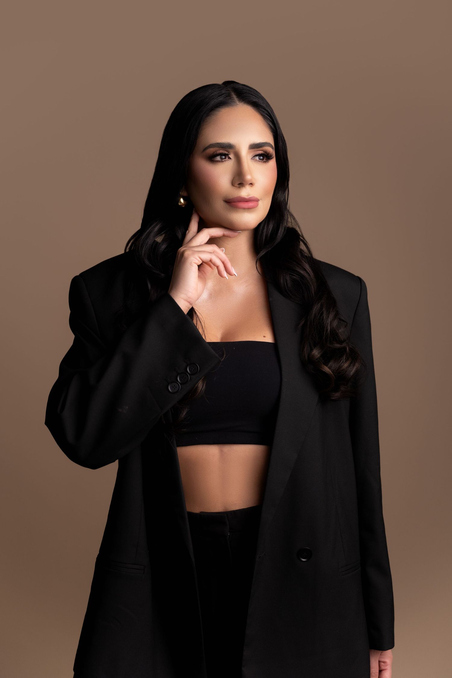Woman in black blazer and crop top poses confidently against a warm brown background with long dark wavy hair.