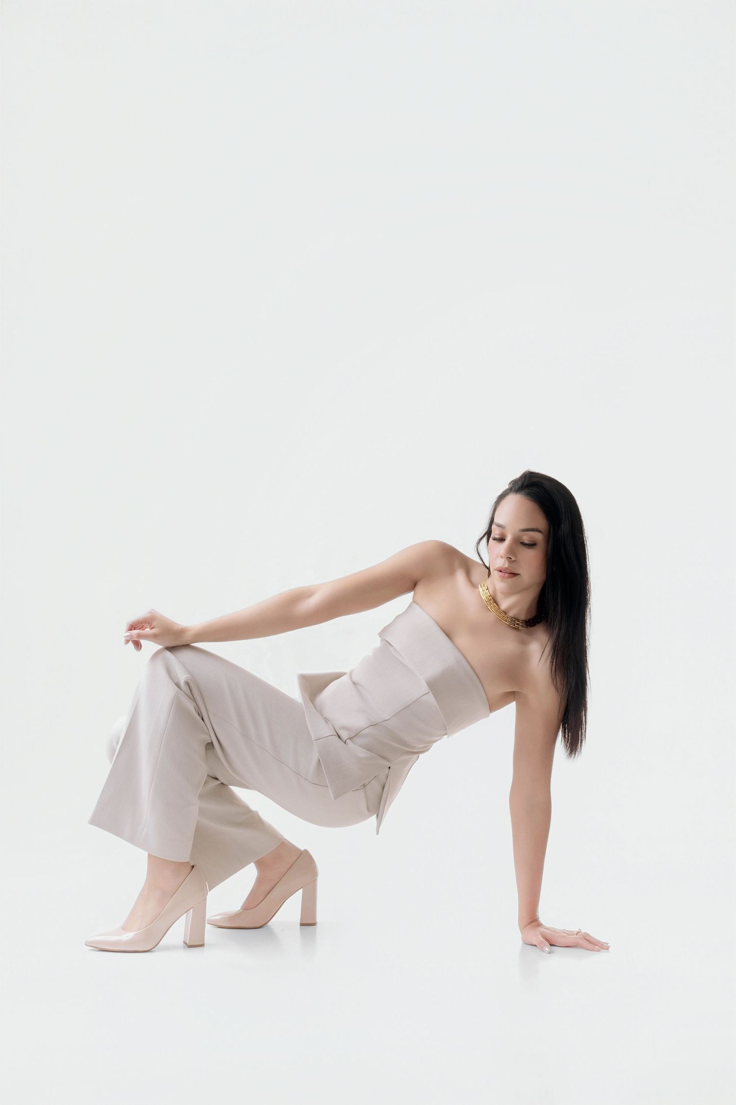 Woman with long dark hair poses low to the ground in a beige strapless jumpsuit and nude pumps against a clean white studio backdrop.