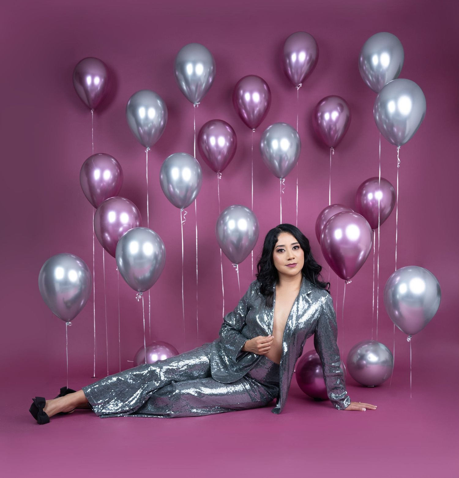 Woman in a sparkling silver sequin suit reclines on a pink backdrop surrounded by metallic silver and pink chrome balloons.